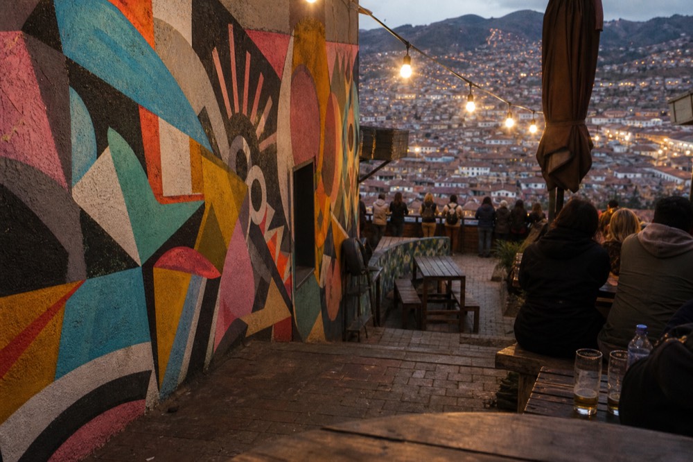 A busy open-air viewpoint terrace above a valley of dense rooftops, visitors seen from behind leaning on a railing, colourful abstract murals on the wall beside them