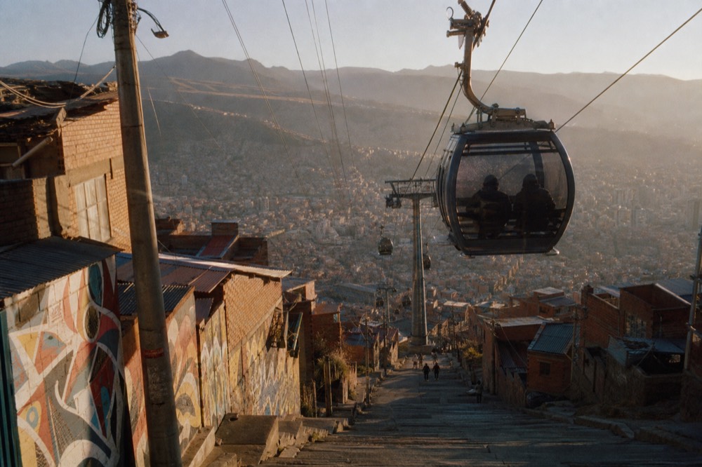 A glass cable-car cabin gliding above a dense hillside of red-brick houses, two passengers seen in silhouette from behind against the valley view