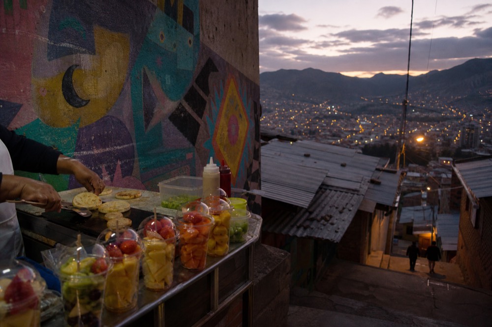 A small street food stall on a hillside landing with fresh fruit cups and a griddle, the vendor's hands in frame, colourful abstract mural wall behind